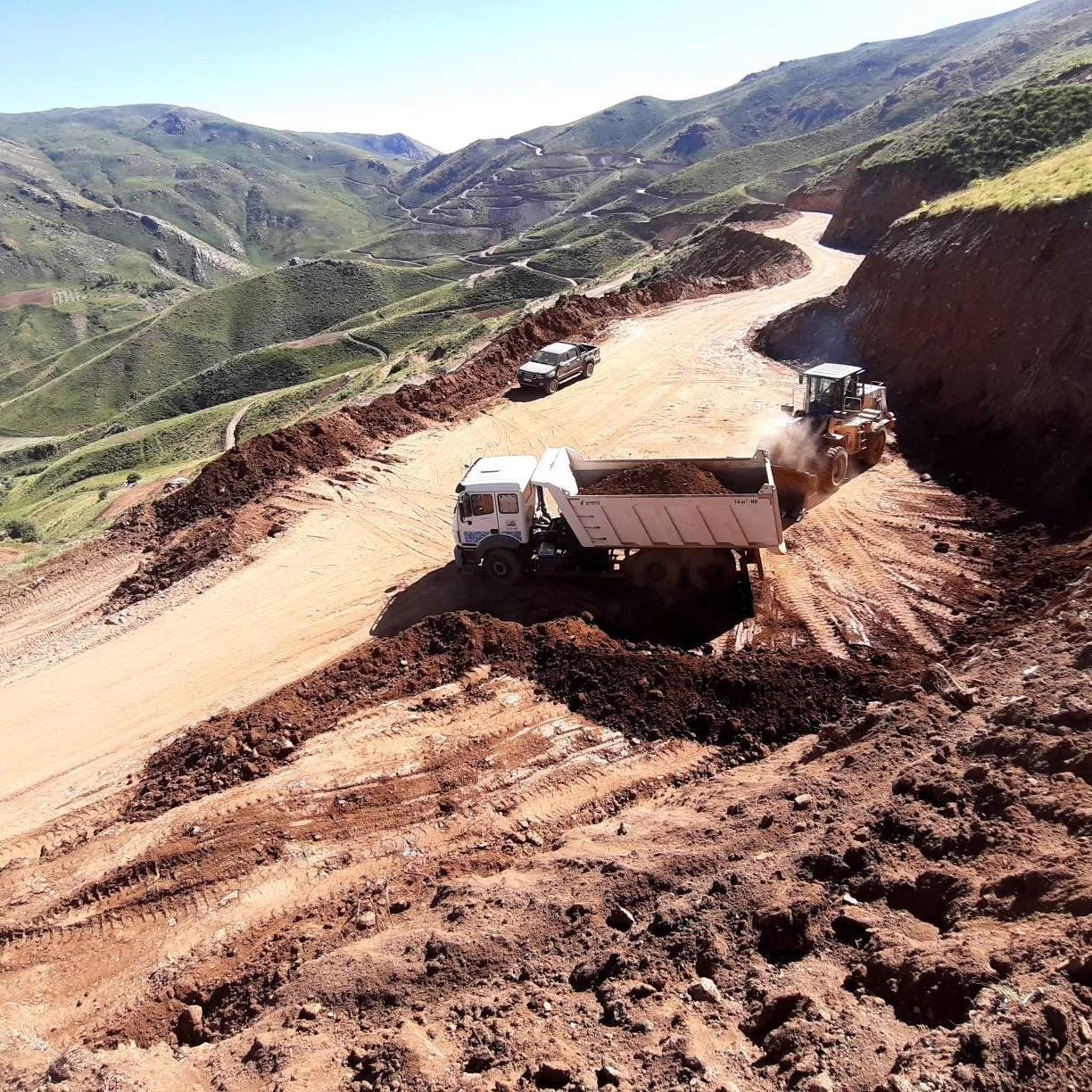 User-supplied field photograph of road-cut earthworks, a dump truck, machinery and a mountain access route